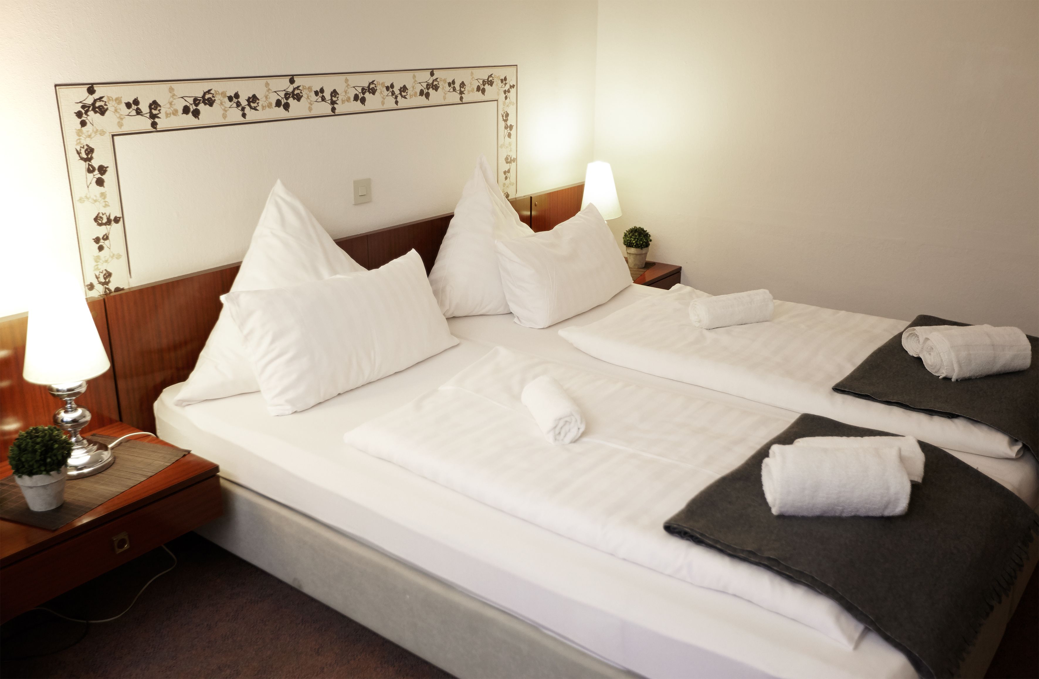 A double bed with white linens, two pillows, a bedside lamp, and a small potted plant.
