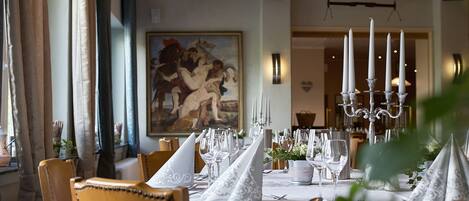 A dining area with a long table set for a meal, featuring white tablecloths, silverware, and candle holders.