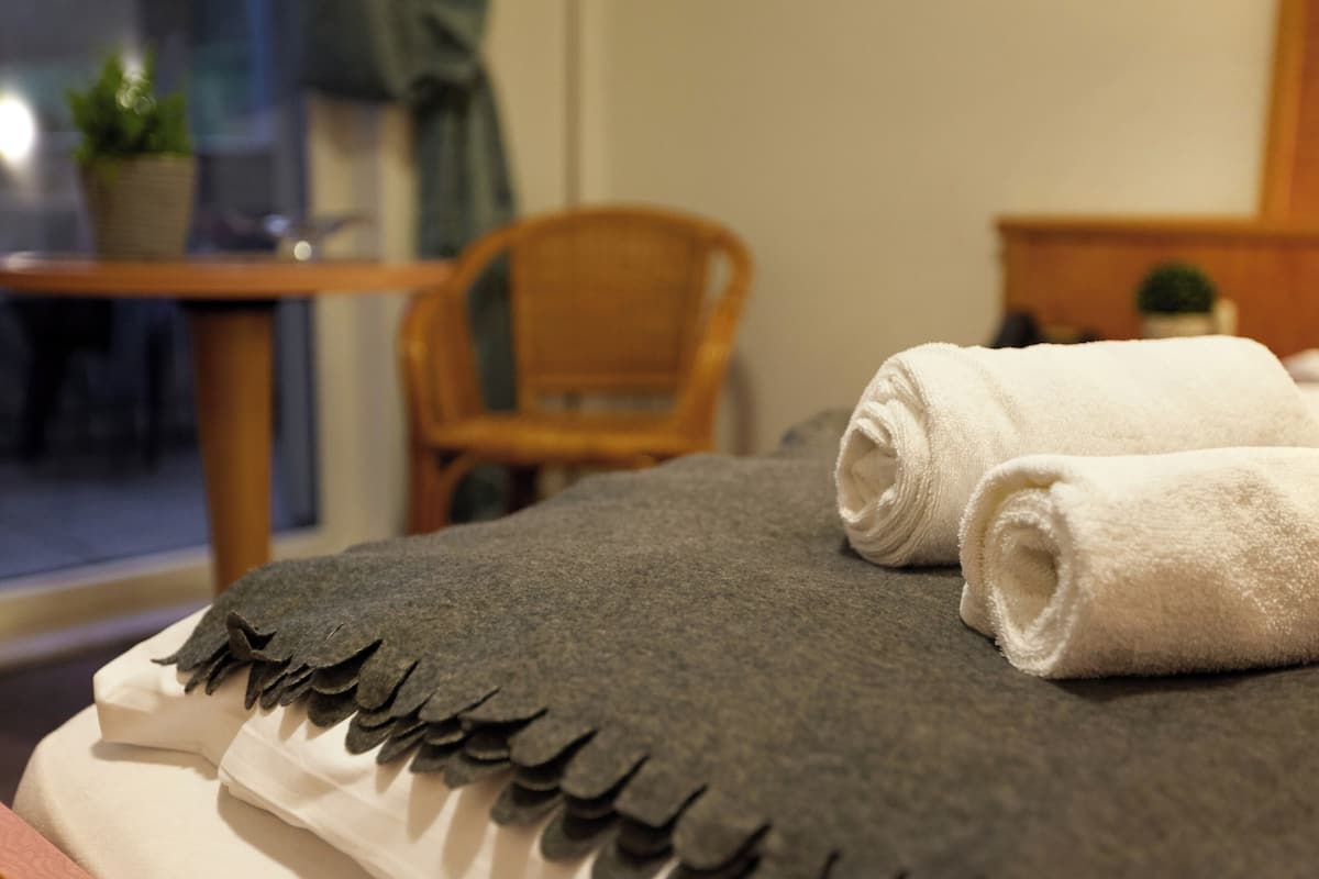 Un lit bien fait, recouvert d’une couverture grise et d’une serviette blanche, une chaise en bois et une petite table sur laquelle est posée une plante.