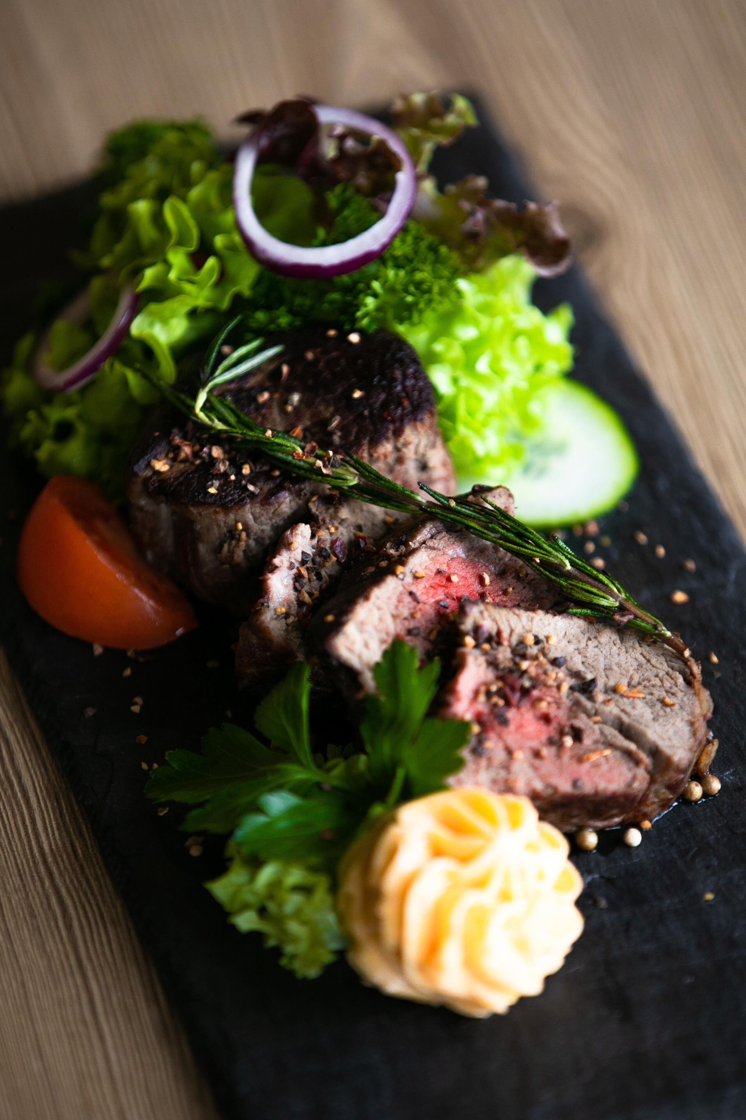 Een steak met een garnering van rozemarijn en peterselie, vergezeld van een salade en een lepel saus op een donker leisteen bord.