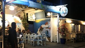 An outdoor dining area with tables and chairs, a sign with a logo, and a building with a blue and white color scheme. - Repag Wayan Canggu Hostel (Canggu)