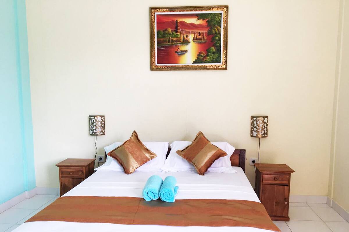 A neatly made bed with two pillows, a blue towel, and a brown blanket. There are two bedside tables with lamps, and a painting of a landscape hangs on the wall.