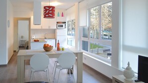 Eine moderne Küche mit weißen Schränken, einem Essbereich mit zwei weißen Stühlen und einem großen Fenster mit Blick auf Bäume und eine Straße.
