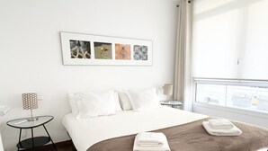 Ein Hotelzimmer mit einem großen Bett, einem kleinen Beistelltisch, einer Lampe und einem Fenster mit Blick auf die Stadt.