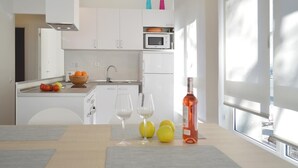Eine moderne Küche mit weißen Schränken, einer Mikrowelle und einer Obstschale. Ein Esstisch mit Platzdeckchen und einer Weinflasche steht daneben.