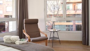 Hotelkamer met een groot bed, een bruine fauteuil, een klein rond tafeltje met een lamp en uitzicht op gebouwen door de ramen.