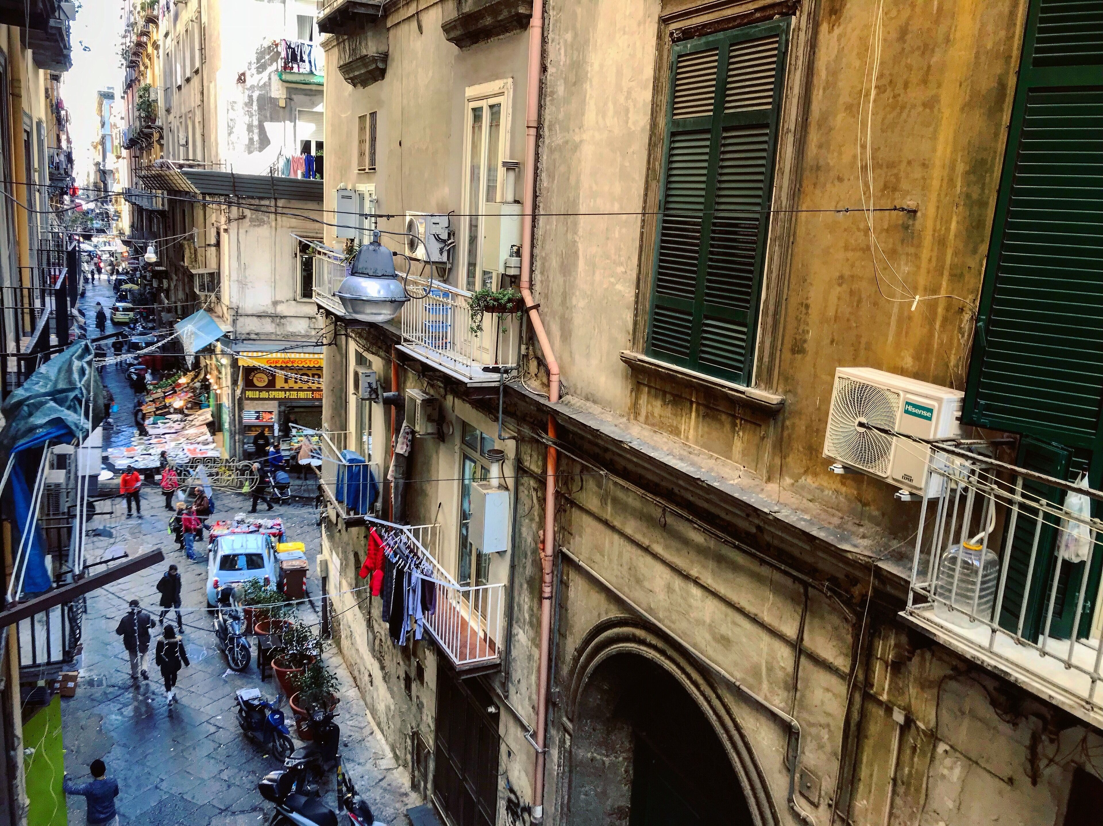 Una calle estrecha flanqueada por edificios, con balcones llenos de ropa tendida y algunas personas caminando.