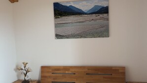 A bedroom with a wooden ceiling, a bed with a white textured cover, a painting of mountains on the wall, and a small plant on a wooden nightstand. - Beautiful apartment in an ideal location for a relaxing, restful vacation (Hausham - Agathried)
