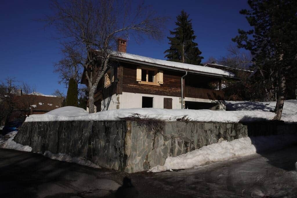 Eine Holzhütte mit schneebedecktem Dach, umgeben von Bäumen und einer Steinmauer.