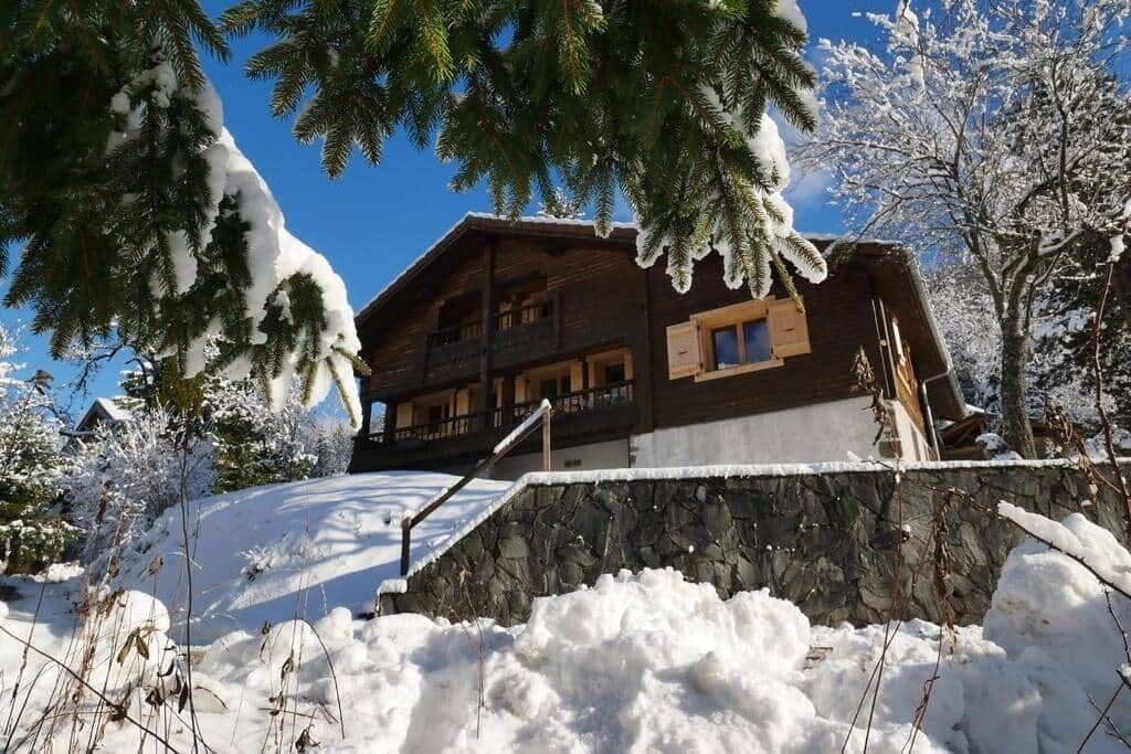 A wooden chalet with a stone wall in front, surrounded by snow-covered trees.