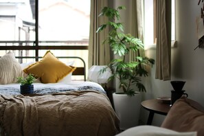 A neatly made bed with a brown blanket, a yellow pillow, and a small potted plant. There is a window with curtains, a lamp, and a small table with a cup and saucer. - Koru Takanawa Gateway Hostel Cafe&Bar (Tokyo)