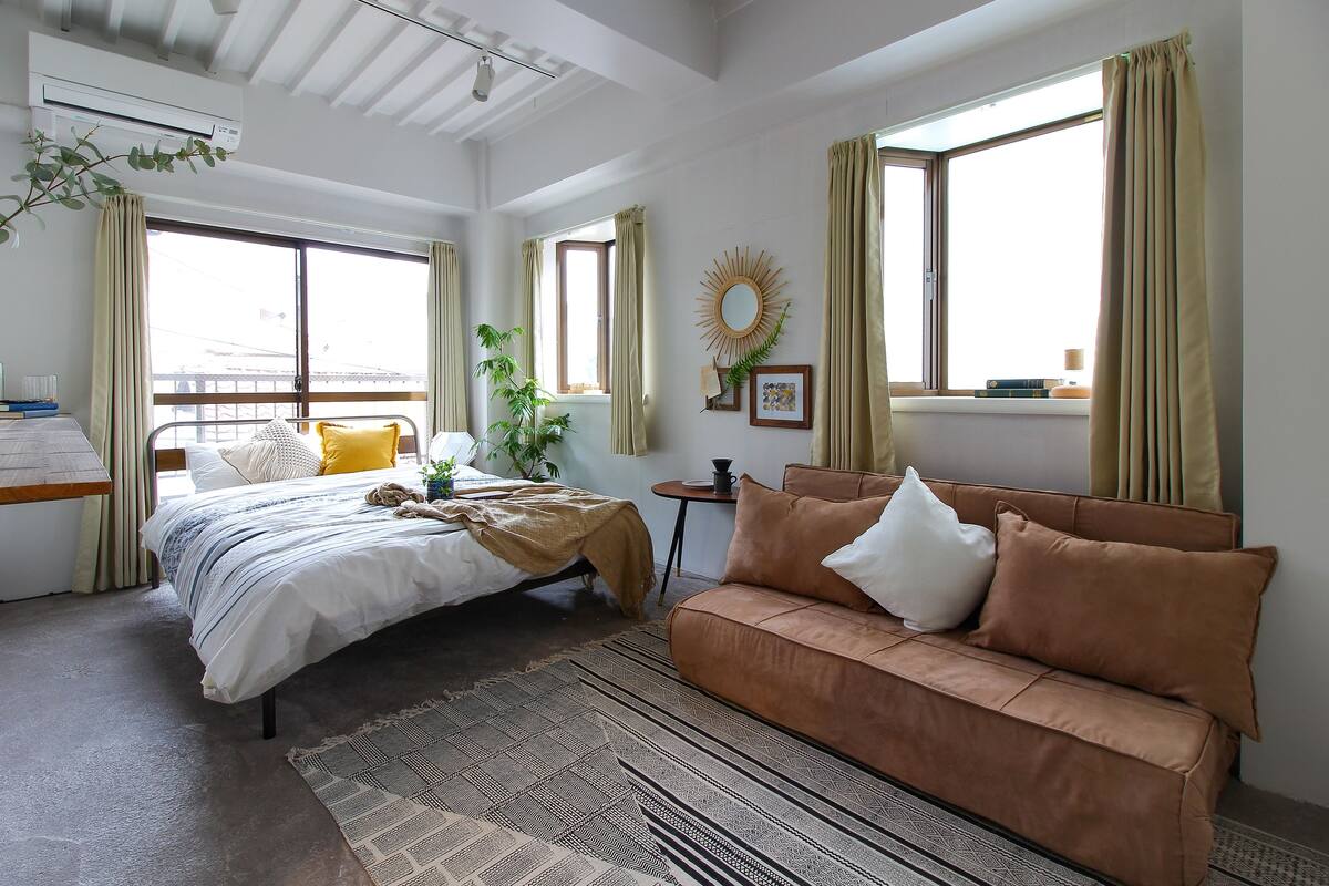 Un dormitorio moderno con cama, sofá marrón, ventana con cortinas, una mesita y un espejo montado en la pared.