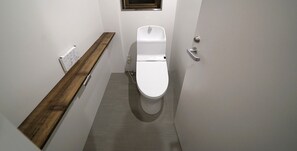 A narrow bathroom with a white toilet, a wooden shelf, and a small window. - Koru Takanawa Gateway Hostel Cafe&Bar (Tokyo)