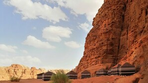A desert landscape with a large rock formation and a row of striped tents. - Wadi Rum Desert Tours Camp (Wadi Rum)