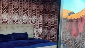 A bedroom with a patterned curtain, a bed with a blue cover, and a view of a mountainous landscape through a window. - Wadi Rum Desert Tours Camp (Wadi Rum)