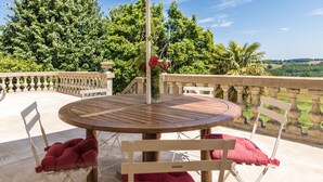 A round wooden table with a white umbrella, surrounded by white chairs with red cushions, set on a balcony with a stone balustrade. - Stylish Mansion in Savignac (Savignac-Ledrier)