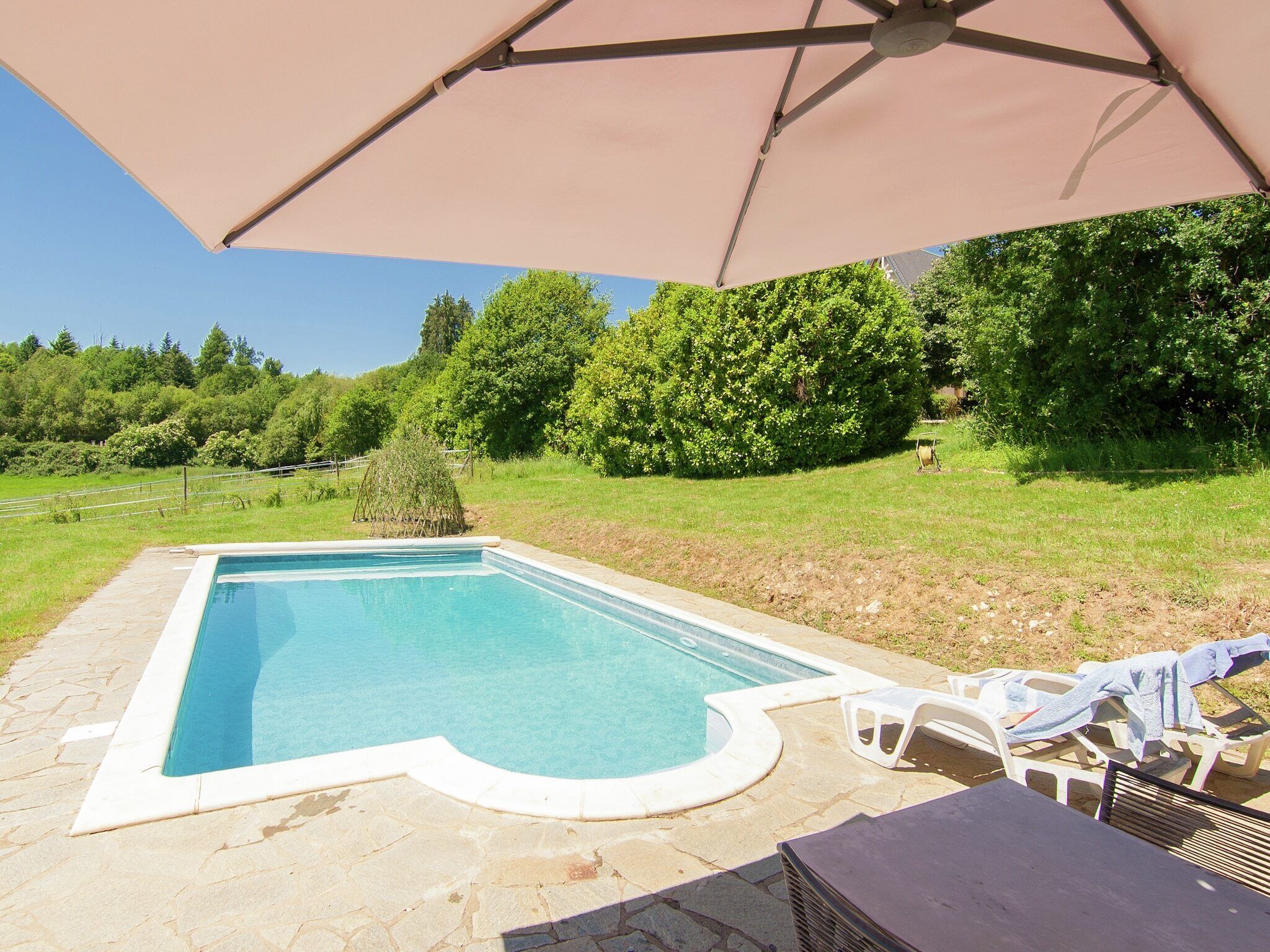 A rectangular swimming pool with a stone border, surrounded by a stone patio. There is a white umbrella providing shade over the pool area.