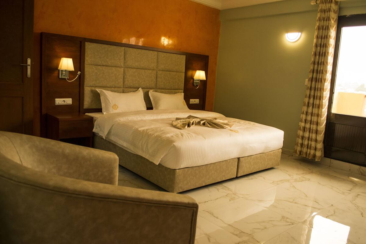 A hotel room with a large bed, two bedside lamps, a nightstand, a chair, and a view of the outside through a window with curtains.