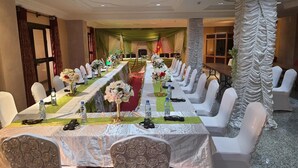 Une salle de conférence avec une longue table préparée pour une réunion, entourée de chaises blanches, agrémentée de compositions florales et de bouteilles d’eau posées sur la table.