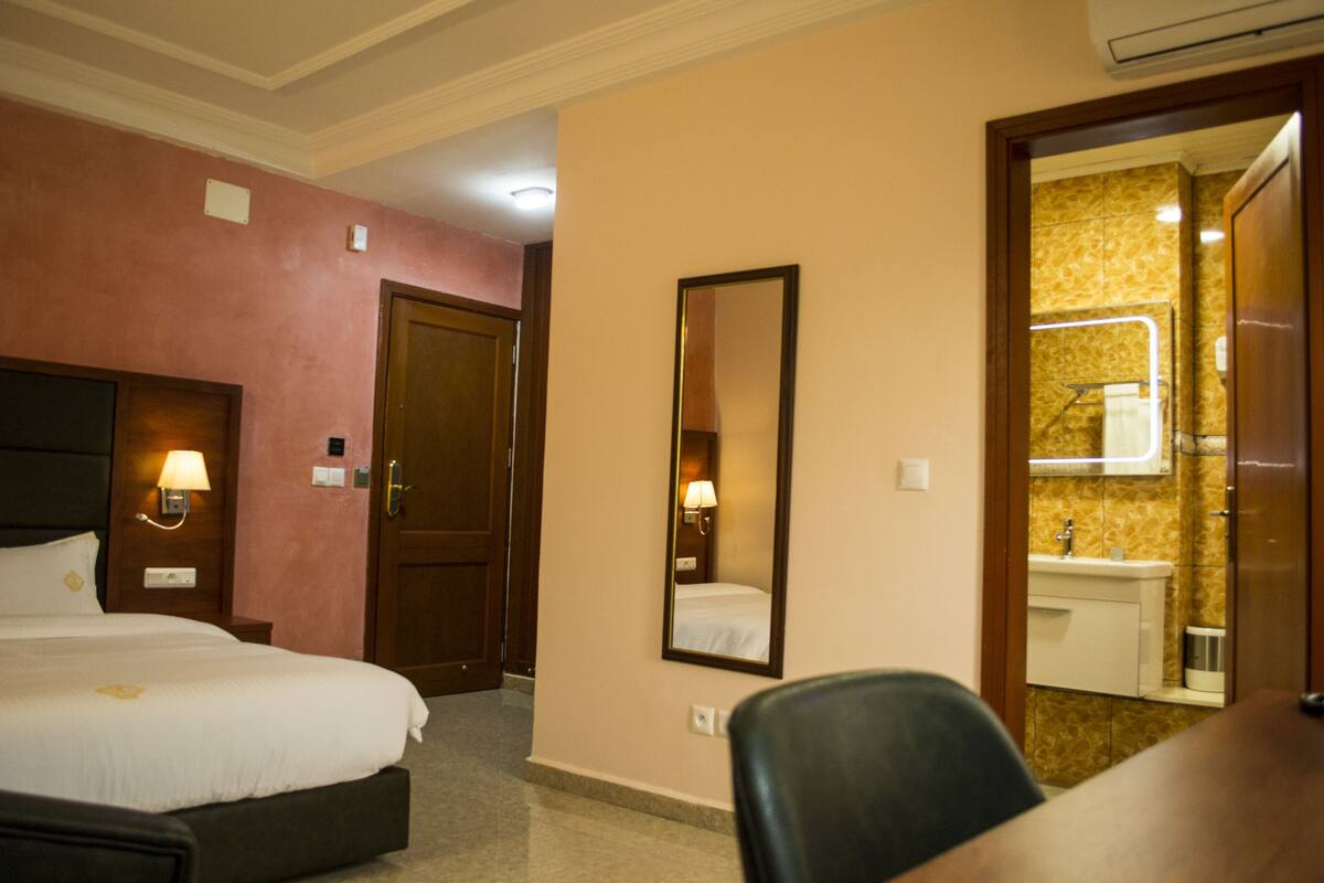 A hotel room with a bed, a desk, a chair, a mirror, and a bathroom visible through an open door.