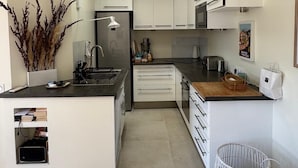 Eine moderne Küche mit weißen Schränken, einer zentralen Kochinsel und einem eingebauten Backofen. Es gibt ein Spülbecken, einen Kühlschrank und eine Mikrowelle. Der Boden ist mit Fliesen belegt und es gibt einen kleinen Essbereich mit Tisch und Stühlen.