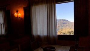 Ein schwach beleuchteter Raum mit einem Holztisch, einem Kerzenständer und einem Fenster mit Blick auf eine gebirgige Landschaft.