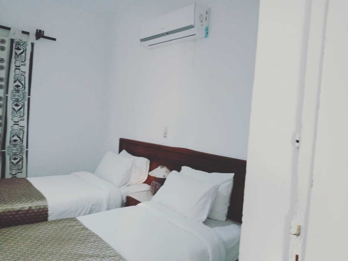 Una habitación con dos camas individuales, cabecera de madera, pared blanca y cortina estampada.