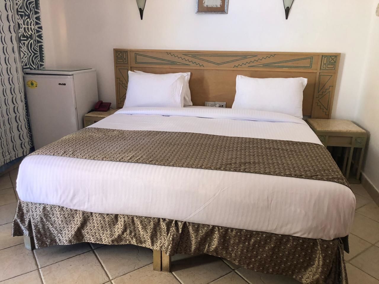 Ein Hotelzimmer mit einem Bett, einem Nachttisch, einem Kühlschrank und einem gemusterten Vorhang.