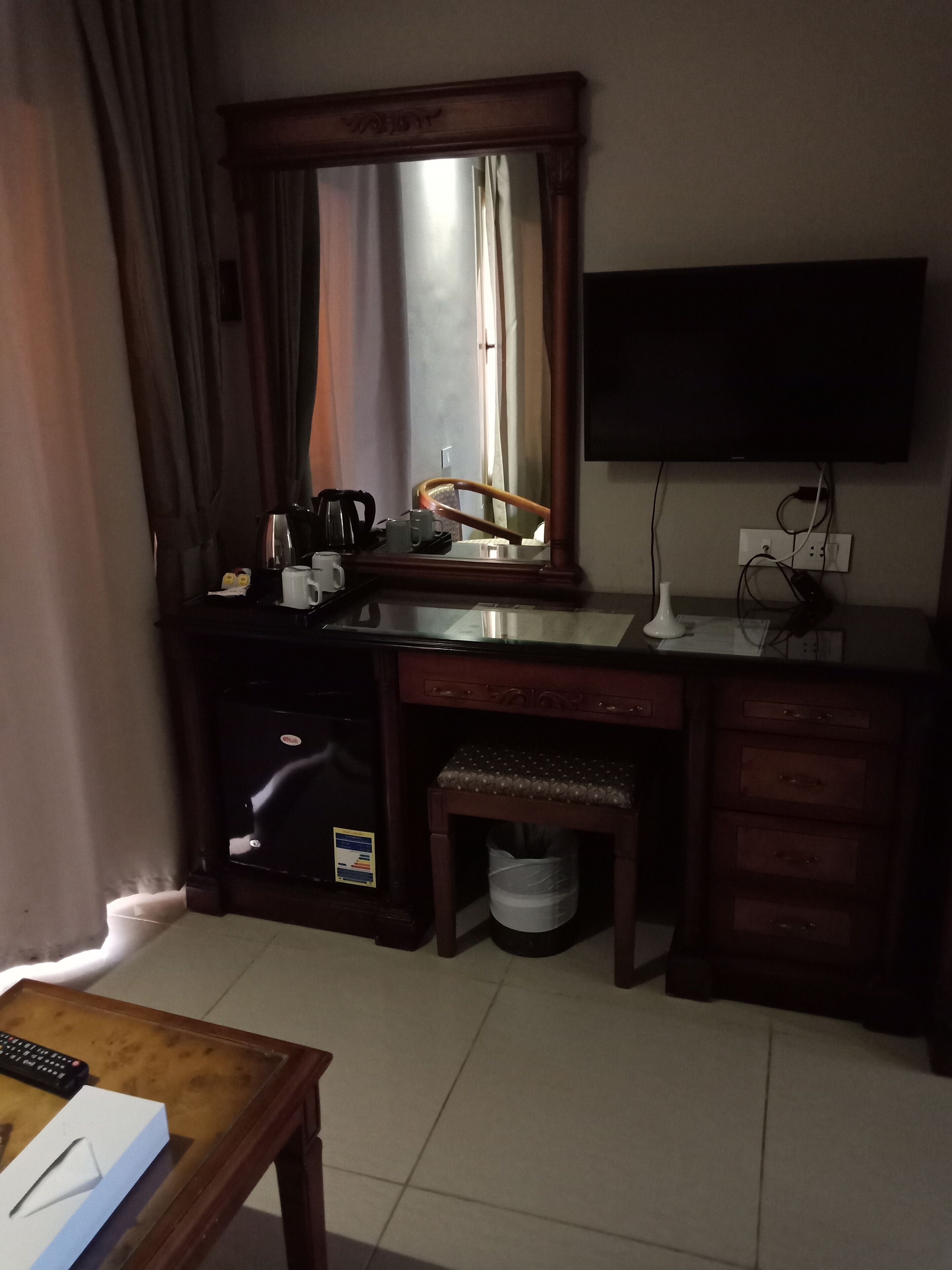 Ein Hotelzimmer mit einem Holztisch, einem Spiegel, einem an der Wand befestigten Fernseher, einem Mülleimer und einem Fenster mit Vorhängen.