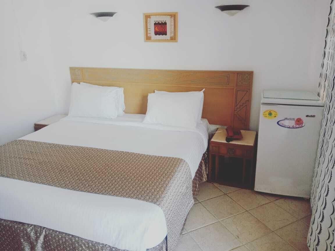 Quarto de hotel com uma cama de solteiro, mesa de cabeceira, pequeno frigorífico e luminária fixada na parede.
