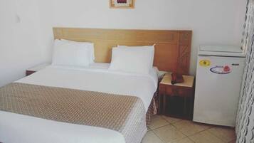 Quarto de hotel com uma cama de solteiro, mesa de cabeceira, pequeno frigorífico e luminária fixada na parede.