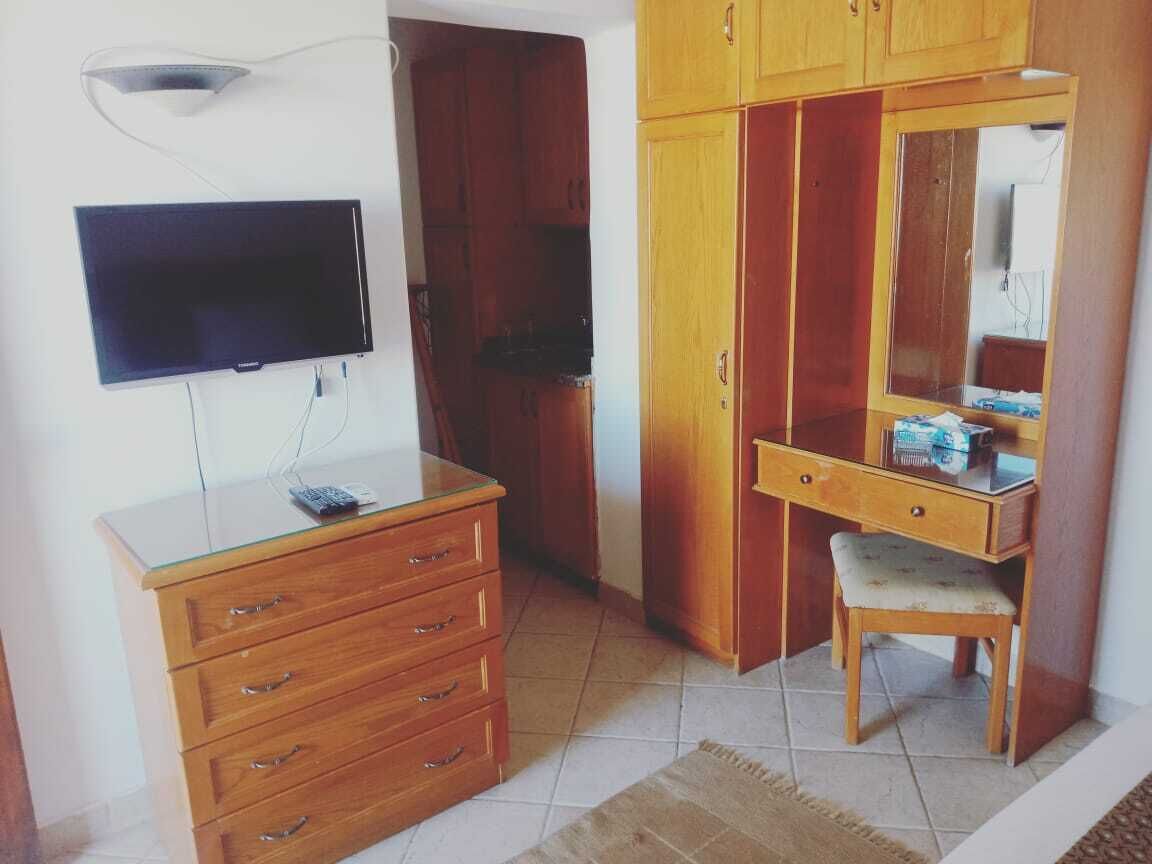 A room with a flat-screen TV mounted on the wall, a wooden dresser with drawers, a desk with a chair, and a mirror cabinet.