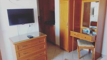 A room with a flat-screen TV mounted on the wall, a wooden dresser with drawers, a desk with a chair, and a mirror cabinet.
