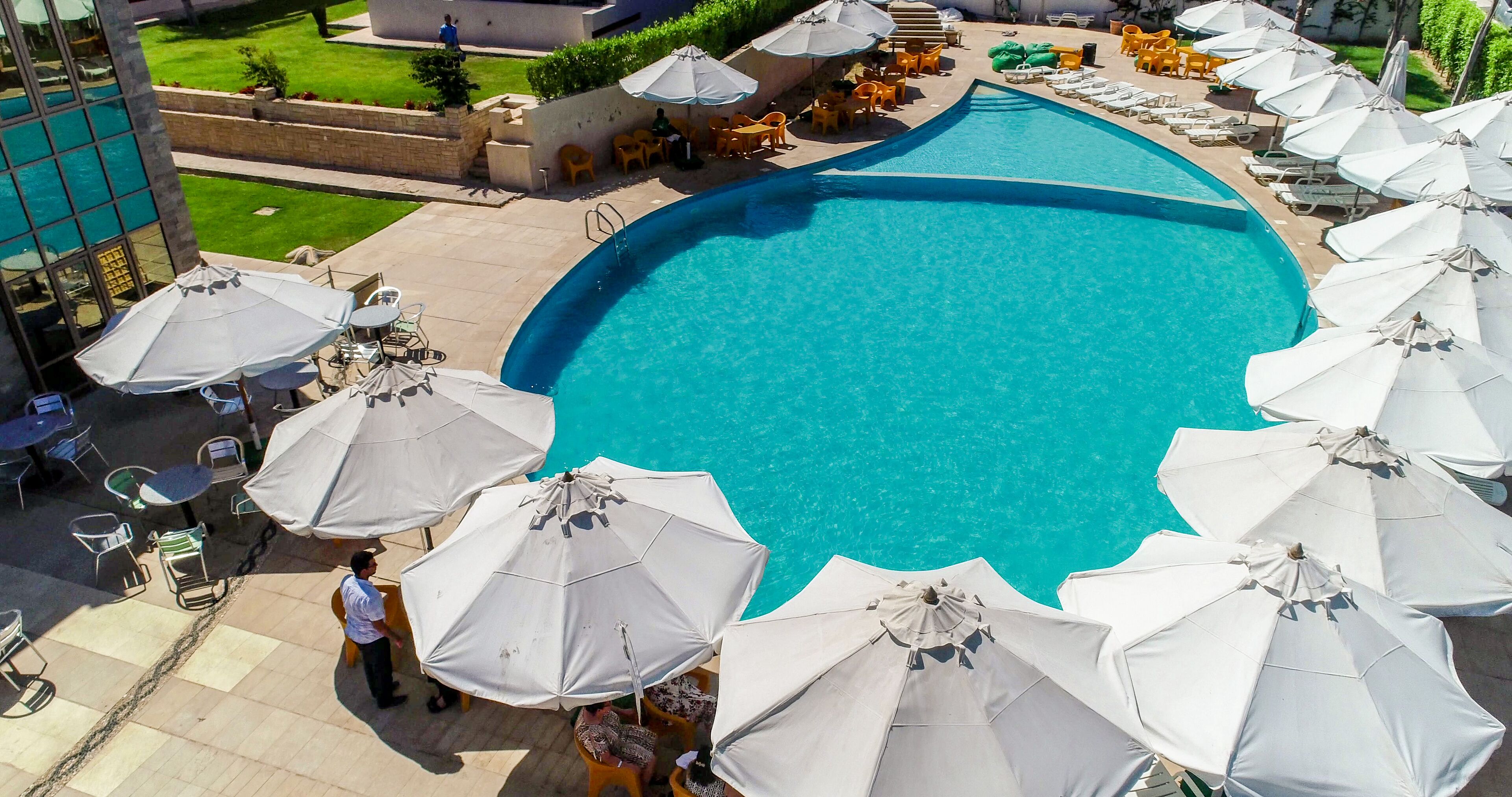 A swimming pool with multiple umbrellas and lounge chairs, surrounded by a landscaped area with steps and a view of a building.