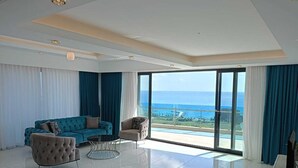 A spacious living room with a large sofa, a round coffee table, and a view of the ocean through floor-to-ceiling windows. - Elite Luxury Suite & Spa (Alanya)