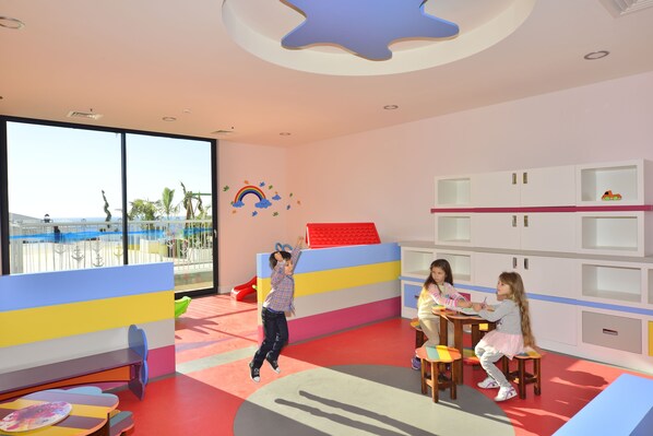 Un cuarto de juegos infantil con muebles coloridos, ventanas amplias y vista a palmeras.