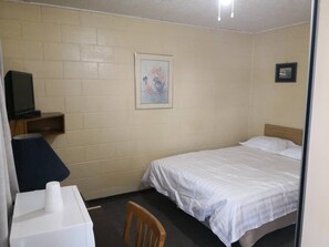 A small, single-bed room with a wooden headboard, a desk with a chair, a television, and a framed picture on the wall.