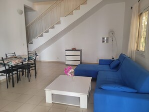 A living room with a staircase, a dining table with chairs, a blue sofa, and a white coffee table with flowers.