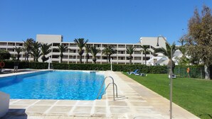 A swimming pool with a clear blue water, surrounded by a paved deck. In the background, there is a multi-story building with balconies and palm trees. - Beautiful apartment by the sea with pool (Roquetas de Mar)