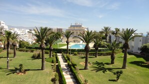 A well-maintained garden with a central pathway leading to a pool area, surrounded by palm trees and modern buildings in the background. - Beautiful apartment by the sea with pool (Roquetas de Mar)