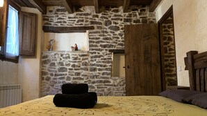 A bedroom with a stone wall, a wooden door, a window with curtains, and a bed with a yellow bedspread.