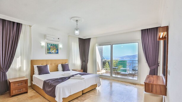 Une chambre spacieuse avec un grand lit, une table de chevet, une petite table en bois et une vue sur un balcon aménagé avec du mobilier d’extérieur.