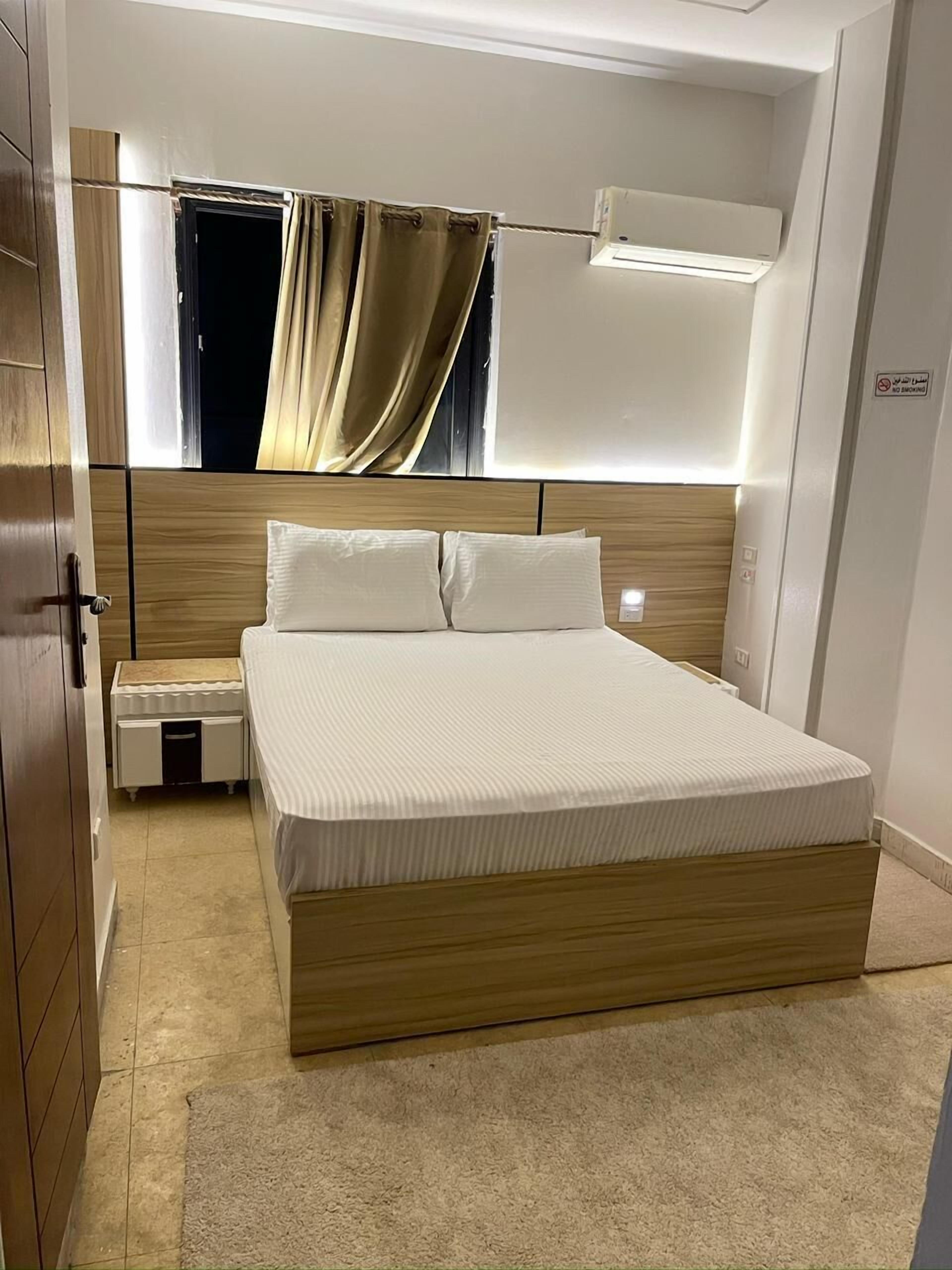 Un dormitorio con cabecera de madera, una cama con ropa blanca, una mesita de noche y una ventana con cortinas.