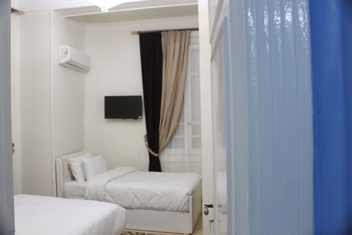 A small, clean room with two beds, a flat-screen TV mounted on the wall, and a window with curtains.