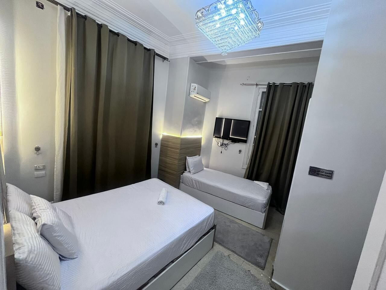 A compact hotel room with two beds, a flat-screen TV, and a chandelier.
