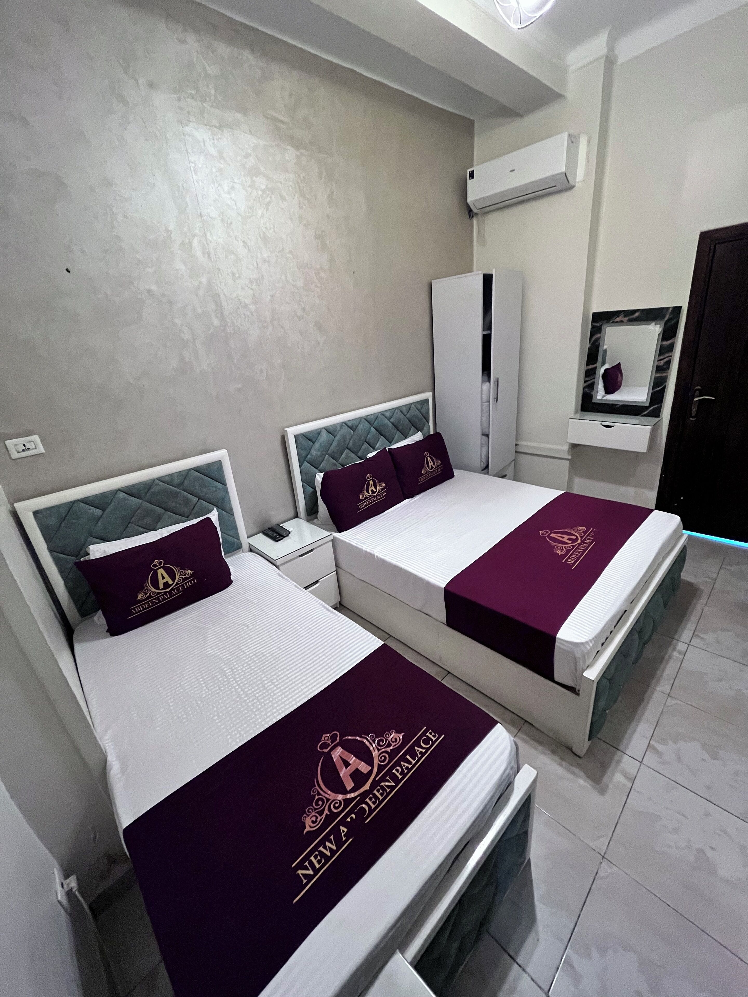 A hotel room with two beds, each with a headboard and a pillow with a logo. There is a wardrobe, a mirror, and an air conditioning unit on the wall.