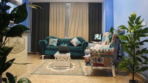 Ein Wohnzimmer mit einem petrolfarbenen Sofa, einem gemusterten Sessel und einem Laptop auf einem Beistelltisch.