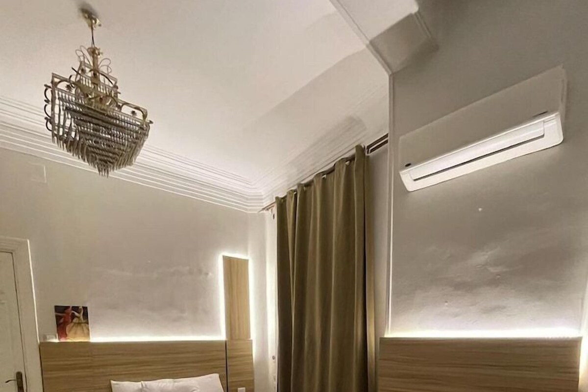 A hotel room with two beds, a chandelier, a bedside table, and a wall-mounted air conditioner.