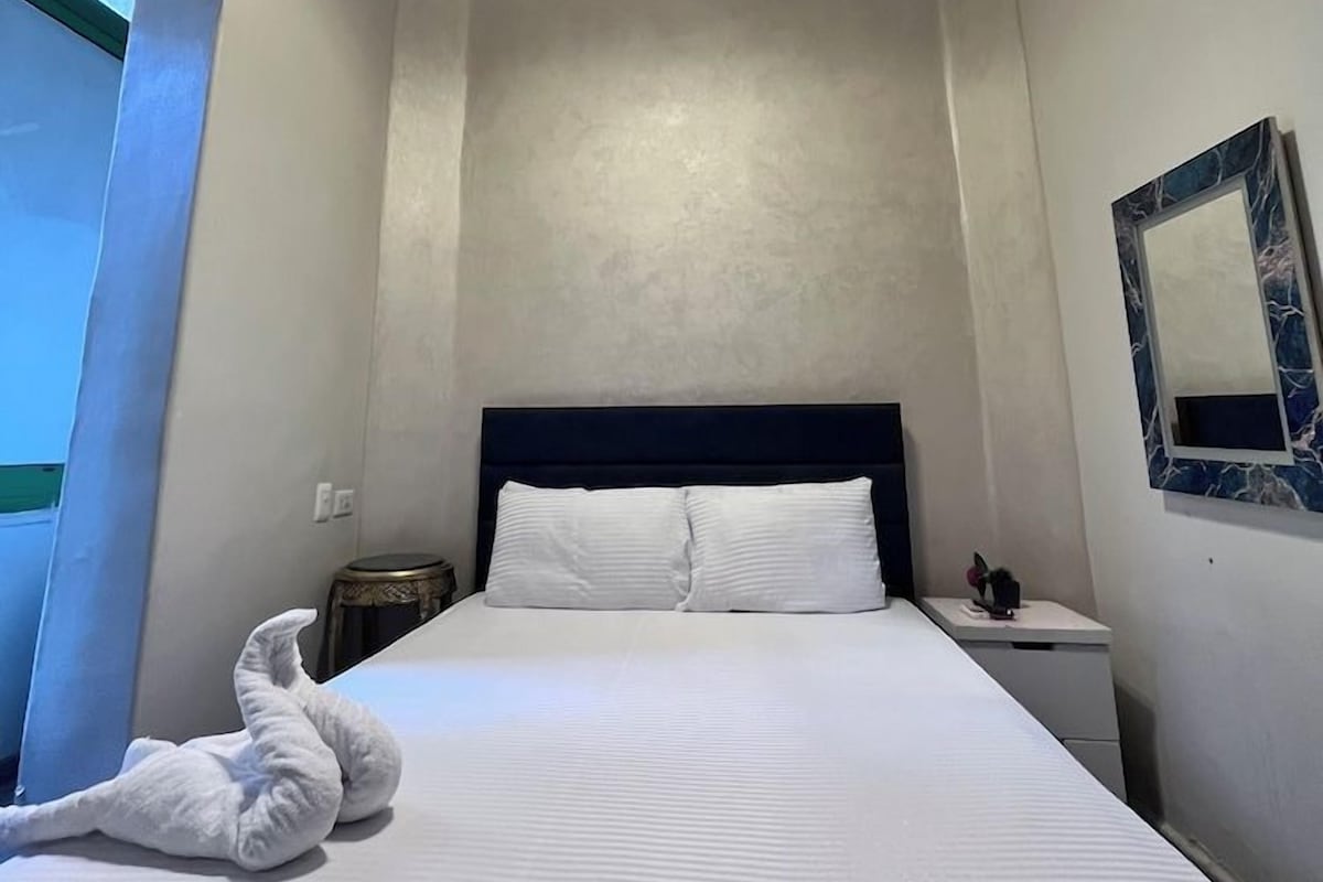 A hotel room with a bed, a bedside table, a wall-mounted mirror, and a ceiling light fixture.