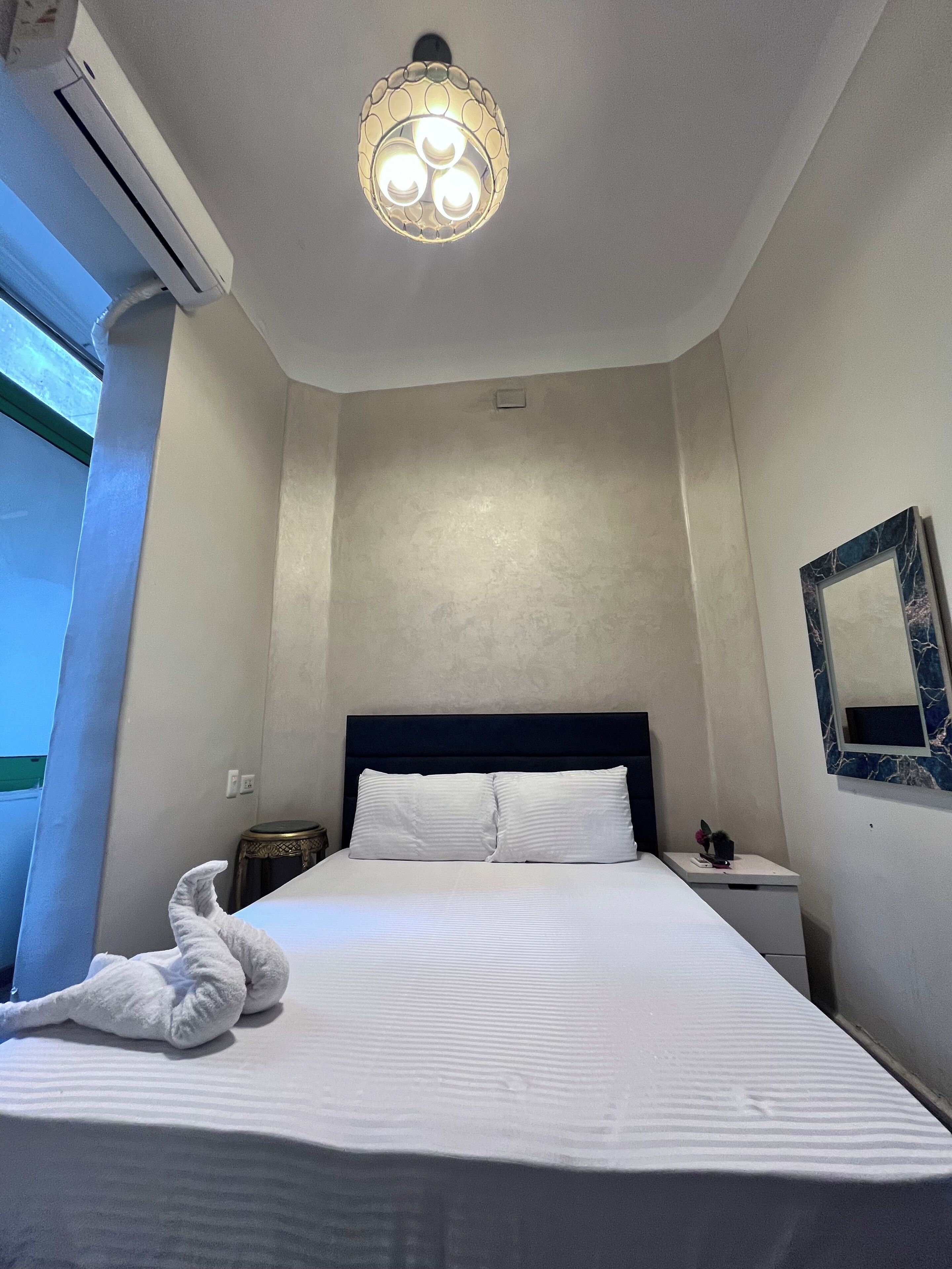 A hotel room with a neatly made bed, a bedside table with a lamp, a wall-mounted mirror, and a ceiling light fixture.